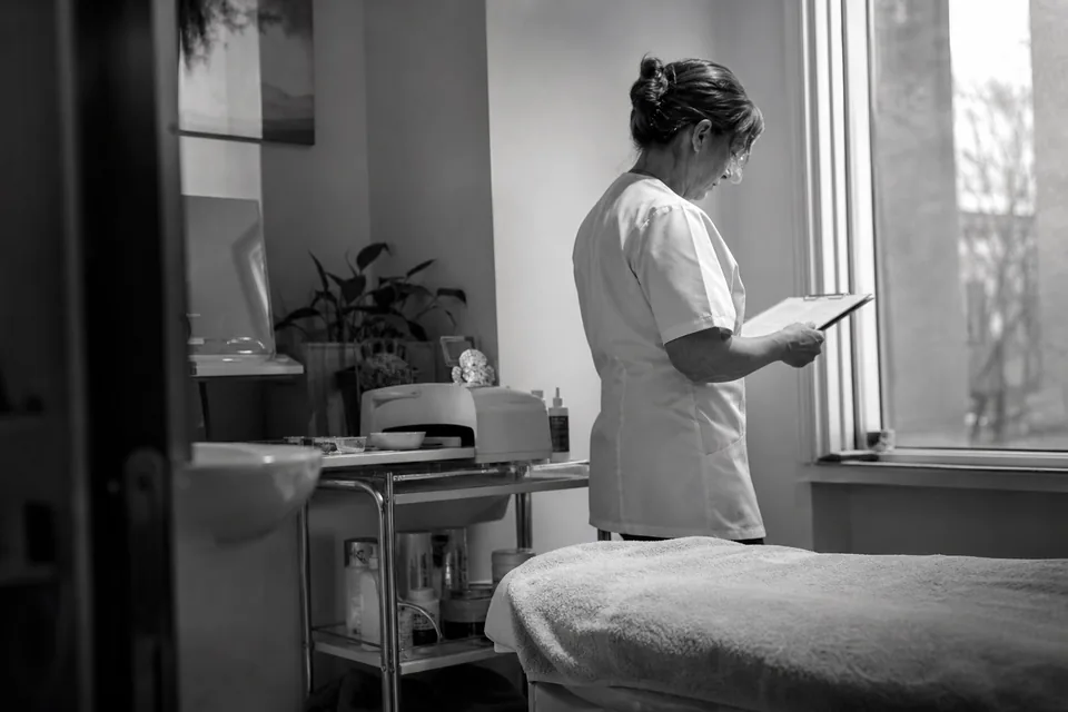 A clinic owner in a white tunic standing in her empty treatment room holding a clipboard