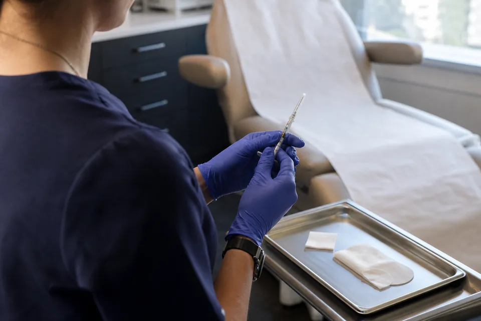 A provider in gloves holding a small sealed filler syringe over a steel tray beside a consultation chair