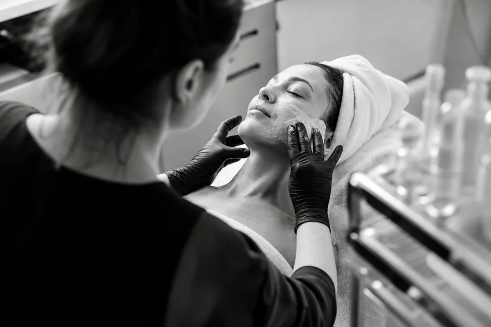 An esthetician's gloved hands smoothing a clay mask on a client's cheek in a bright treatment room