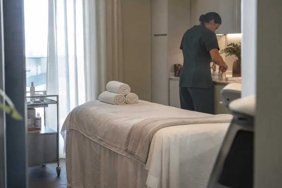 An empty treatment room with a linen-covered bed, folded towels and a window with sheer curtains