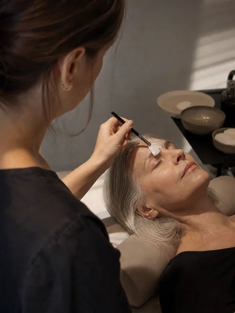 An esthetician applying a serum with a brush to a reclined client in a sunlit studio